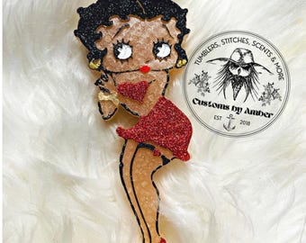 May include: A glittery resin figure of a cartoon character with black hair, a red dress, and red shoes, holding a red heart. Next to the figure is a circular logo with the text TUMBLERS, STITCHES, SCENTS & MORE, Customs by Amber, EST 2018.