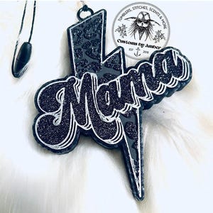 May include: A black lightning bolt-shaped car air freshener featuring a glittery leopard print pattern and the word Mama in a cursive script. The text is outlined in silver, and a black hanging cord is attached to the top loop. A circular logo sticker reads Tumblers, Stitches, Scents & More Customs by Amber Est 2018.