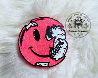 May include: A circular hot pink glittery object featuring a black smiley face design where the right side is replaced by a white skeletal jaw and skull detail. A circular logo with the text Tumblers, Stitches, Scents & More Customs by Amber Est 2018.