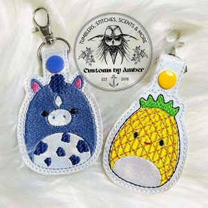 May include: Two embroidered keychains, one with a blue and white cartoon horse and the other with a yellow cartoon pineapple. Both keychains have a white background and a white border. The keychains are on a white fluffy surface. The text on the keychain says "Tumblers, Stitches, Scents & More Customs by Amber Est 2018."