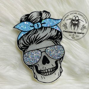 May include: A skull-shaped ornament featuring a messy bun hairstyle with a light blue glitter headband and silver glitter sunglasses. A circular logo is visible in the upper right corner with the text Tumblers, Stitches, Scents & More, Customs by Amber, Est 2018.