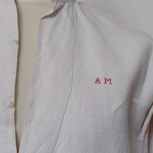 May include: A white linen shirt with a mandarin collar and a red embroidered monogram "A M" on the chest.