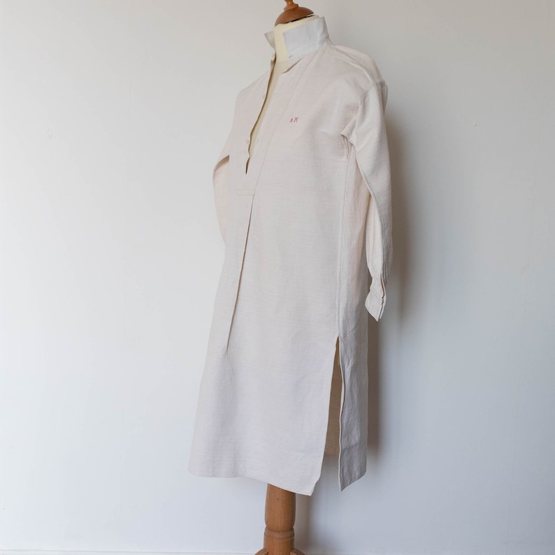 May include: A long-sleeved, off-white linen shirt with a V-neck and a side slit. The shirt has a small embroidered monogram on the chest that reads "A.P."