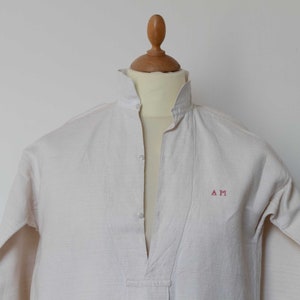 May include: A white linen shirt with a mandarin collar and a button-up front. The shirt has a small embroidered monogram on the chest that reads "A M".