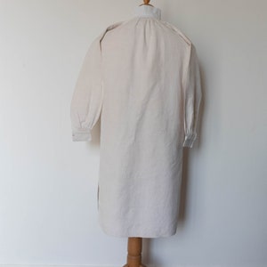 May include: A long-sleeved, white linen shirt with a gathered neckline and buttoned cuffs. The shirt is displayed on a mannequin.