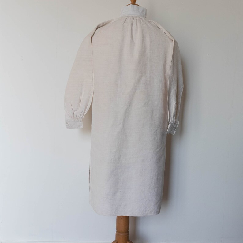 May include: A long-sleeved, white linen shirt with a gathered neckline and buttoned cuffs. The shirt is displayed on a mannequin.
