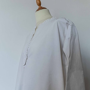 Può includere: Camicia bianca in cotone con un bordo di pizzo intorno al collo. La camicia ha maniche lunghe e una vestibilità rilassata.
