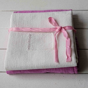 May include: Two white linen tea towels with a pink ribbon tied around them. The towels have a small embroidered square in the center.