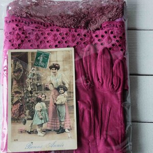 Può includere: Un biglietto postale vintage con una scena natalizia con tre persone, un albero decorato e il testo "Bonne Année". Il biglietto postale è confezionato con un paio di guanti rosa.