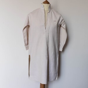 May include: A long-sleeved, white linen shirt with a mandarin collar and a single button closure at the neck. The shirt has a split side seam and the initials "A.M." embroidered on the chest.