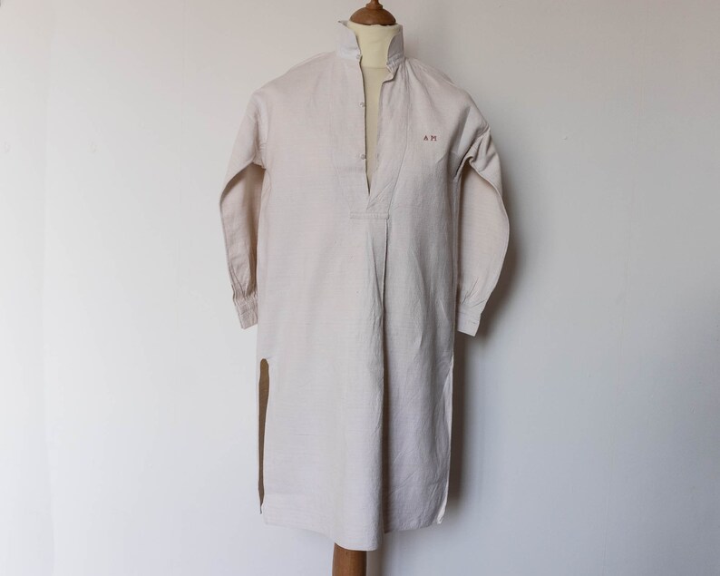 May include: A long-sleeved, white linen shirt with a mandarin collar and a single button closure at the neck. The shirt has a split side seam and the initials "A.M." embroidered on the chest.