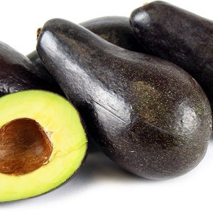 May include: A group of ripe avocados, with one cut open to reveal the green flesh and large brown seed. The avocados have dark, almost black, skin and are arranged on a white background.