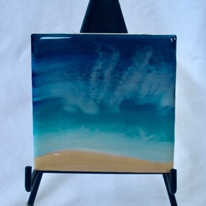 May include: A small resin art piece depicting a beach scene with blue and turquoise water, white foam, and a sandy beach. The art is displayed on a black easel.