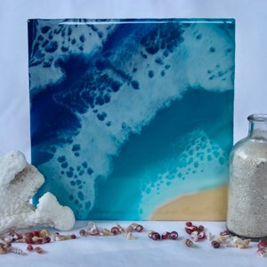 Könnte beinhalten: Ein Harzbild, das einen blauen Ozean mit weißem Schaum und einen Sandstrand darstellt. Das Bild ist gerahmt und auf einer weißen Oberfläche mit Muscheln und einer Glasflasche gefüllt mit Sand ausgestellt.