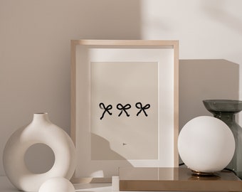 May include: A rectangular light wood frame containing a beige print with three black hand-drawn style bows arranged horizontally in the center. Below the bows, a small black symbol is visible. The frame is positioned upright on a flat surface.