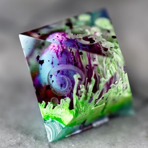 May include: A multi-coloured, translucent, geometric resin object. The object features swirls of purple, green, and blue, with black speckles. The design includes a spiral pattern and a textured, organic appearance. The object is set against a grey background.