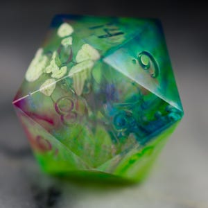 May include: A green, blue, and pink iridescent resin dice with white flowers and numbers embedded inside. The dice is a D6, with the number 9 visible on one side.