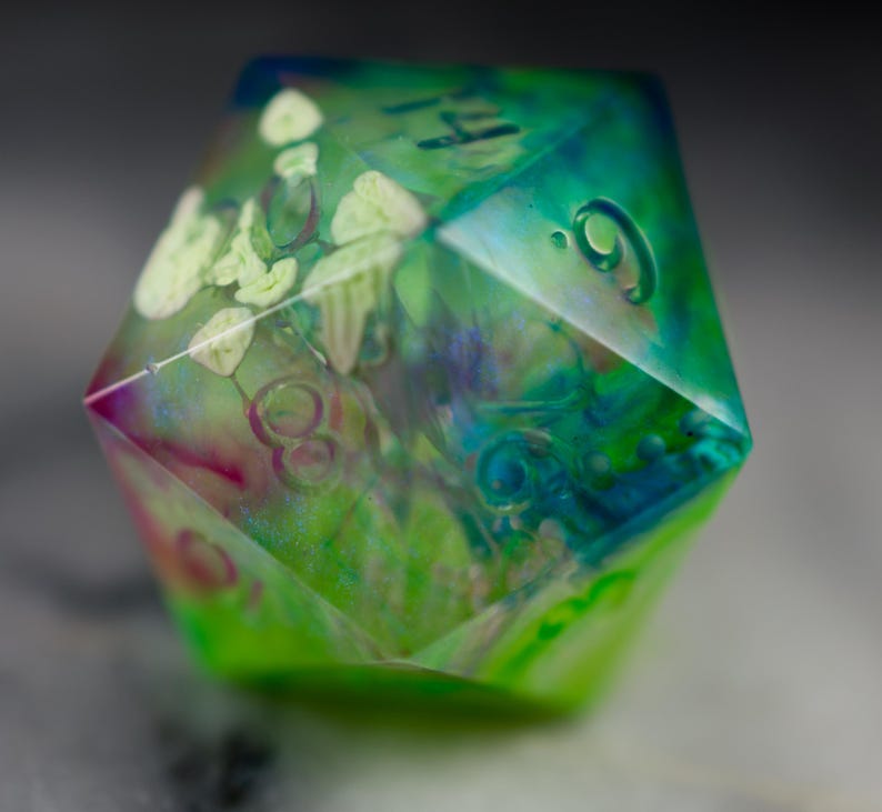 May include: A green, blue, and pink iridescent resin dice with white flowers and numbers embedded inside. The dice is a D6, with the number 9 visible on one side.