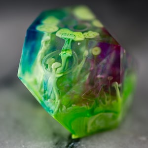 May include: A green, blue, and purple iridescent dice with a white mushroom design inside. The dice is a polyhedral shape with many sides.
