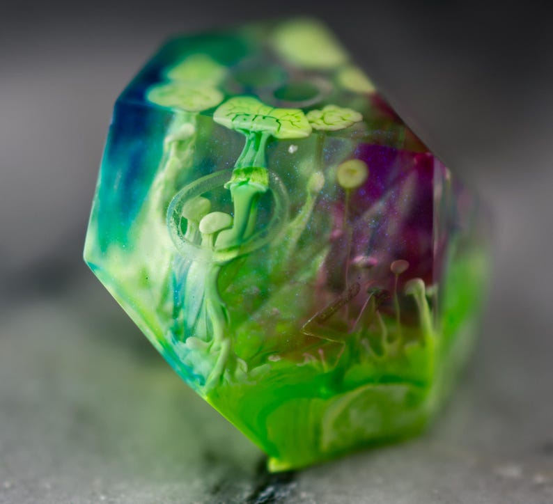 May include: A green, blue, and purple iridescent dice with a white mushroom design inside. The dice is a polyhedral shape with many sides.