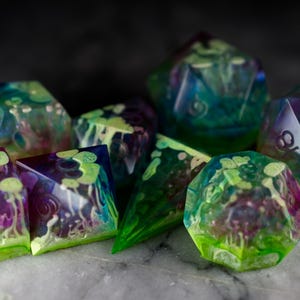 May include: A set of seven iridescent dice with a green, blue, and purple swirl pattern. The dice are made of clear resin and have a translucent appearance. The dice are perfect for tabletop gaming.