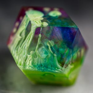 May include: A multi-faceted, iridescent green, blue, and purple resin dice with white swirls and a green base.
