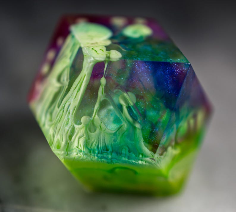May include: A multi-faceted, iridescent green, blue, and purple resin dice with white swirls and a green base.