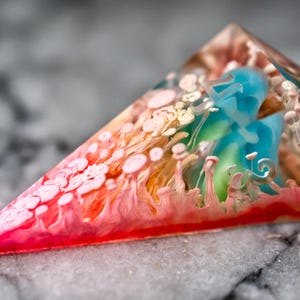 Puede incluir: Un prisma triangular de resina con un exterior transparente que encierra patrones internos arremolinados de pigmentos rojo vibrante, rosa suave y azul brillante. El diseño incluye formas orgánicas similares a nubes y degradados de color distintos.