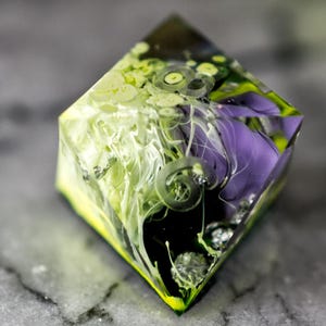 May include: A clear, diamond-shaped resin object with a vibrant mix of colours. The interior features swirls of white and green, with accents of purple and yellow. The design includes spiral shapes and small, reflective elements.