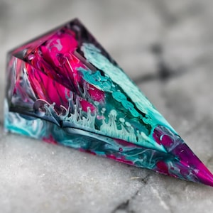 Puede incluir: Un objeto con forma de prisma triangular hecho de resina translúcida que presenta patrones de remolinos en colores magenta vibrante, negro profundo y azul cerceta. El diseño interno muestra vetas fluidas y marmoladas.
