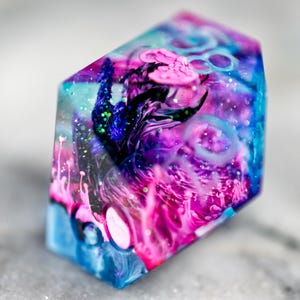 Peut inclure: Objet en résine transparent, à multiples facettes, avec un motif interne tourbillonnant de couleurs rose, violet, bleu et turquoise. Il présente des paillettes bleu foncé et vertes, évoquant une galaxie. Objet décoratif.