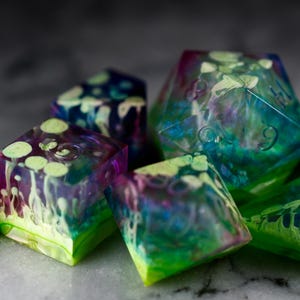 May include: A set of iridescent dice with green, blue, and purple swirls. The dice are made of clear resin and have a translucent appearance.