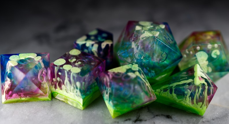 May include: A set of iridescent dice with green, blue, and purple swirls. The dice are made of clear resin and have a translucent appearance.