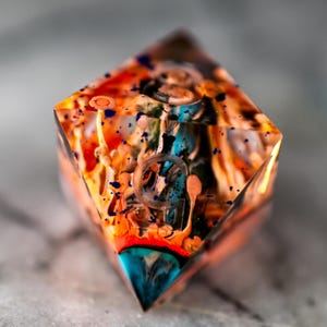 May include: A multi-coloured, transparent resin die with a vibrant, abstract design. The die features a mix of orange, blue, and white hues, creating a unique, artistic appearance. The die is a D20, a twenty-sided die, commonly used in tabletop games.