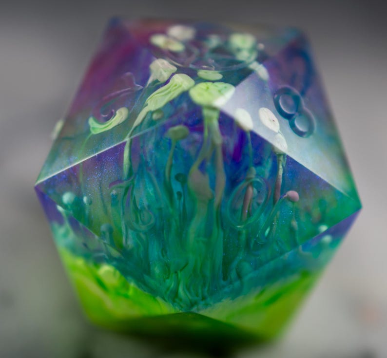 May include: A multi-faceted, clear resin dice with swirls of blue, green, and purple paint. The dice has a white, green, and blue abstract design inside.
