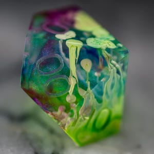 May include: A multi-colored, iridescent, resin dice with green, blue, purple, and white swirls and patterns. The dice is a geometric shape with multiple sides.