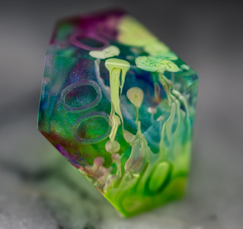 May include: A multi-colored, iridescent, resin dice with green, blue, purple, and white swirls and patterns. The dice is a geometric shape with multiple sides.