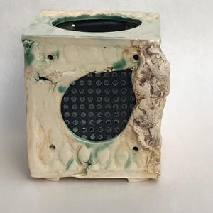 Peut inclure: Un pot de fleurs carré en céramique faite à la main, avec une surface texturée blanc cassé. Le pot a une ouverture circulaire avec un écran en maille noire et des trous décoratifs. Des accents verts et bruns ajoutent de la couleur.