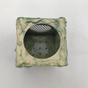 May include: Square ceramic planter with a textured, off-white and green exterior. The planter has a circular opening with a wire mesh and several drainage holes at the bottom. The design is rustic and handmade.