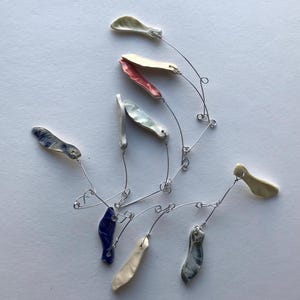 May include: A mobile featuring multiple small, elongated ceramic pieces in various colors including cream, coral, blue, and grey. The ceramic elements are suspended from thin, silver-toned metal wires that are connected with small loops and hooks.