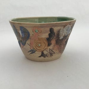 Peut inclure: Un bol en céramique fait à la main avec un extérieur brun clair et un intérieur vert. Le bol est décoré de motifs floraux et abstraits colorés en bleu, rose et noir. Le bol a une forme légèrement évasée.