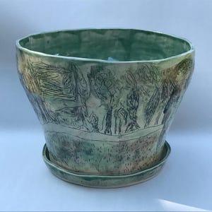 May include: A handcrafted ceramic planter with a light green glaze. The exterior features a detailed, etched landscape scene with trees and figures. Includes a matching saucer for drainage. A unique piece for home decor.