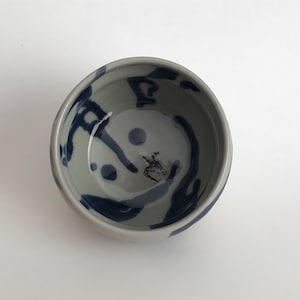 Puede incluir: Un pequeño cuenco de cerámica blanca con un diseño abstracto azul y gris. El cuenco está esmaltado y tiene un acabado liso y brillante.