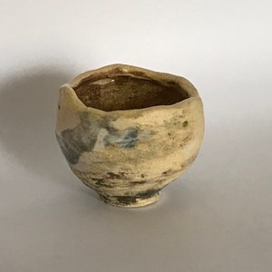 May include: A small, handmade ceramic bowl with a rustic, organic aesthetic. The bowl has a cream-coloured base with mottled grey and blue accents. The interior is a darker brown, and the rim has an uneven, textured edge. The bowl has a small, rounded base.