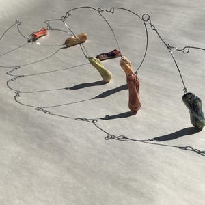 May include: A handmade mobile sculpture featuring a series of colorful, organic-shaped ceramic pieces suspended by thin, silver-colored wire. The ceramic elements are in shades of white, beige, orange, green, and maroon. The mobile casts shadows on the white surface.