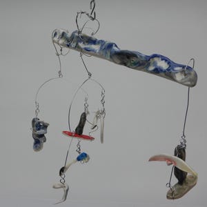 May include: A hanging mobile featuring a long, marbled blue and white ceramic bar suspended by wire. Attached to the bar are several thin wire arms holding various small, abstract ceramic shapes in shades of blue, white, red, and grey.