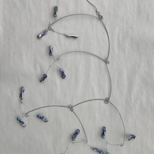 May include: A delicate necklace featuring a silver-toned wire design with multiple curved sections. Small, oblong ceramic beads with blue and white patterns are suspended from the wire, creating a unique and artistic aesthetic. The necklace has a simple chain.