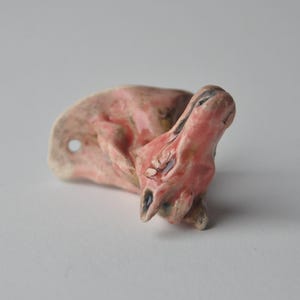 Puede incluir: Una pequeña figura de cerámica esculpida a mano con forma de cabeza de caballo y un esmalte rosa pálido brillante. La pieza presenta una forma suave y redondeada con sutiles hendiduras y un pequeño orificio circular en el lateral.