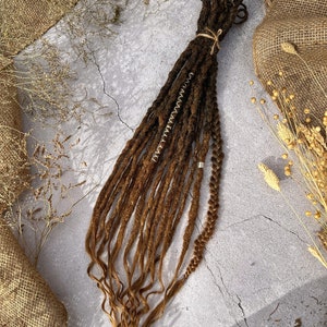 Puede incluir: Un paquete de rastas sintéticas ombre marrón y rubio con cuentas doradas. Las rastas están dispuestas en una pila suelta sobre una superficie gris claro.