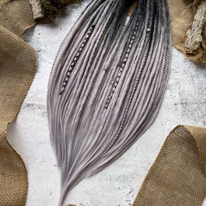 Puede incluir: Un conjunto de rastas sintéticas ombre con un degradado de color de negro a gris claro. Las rastas están dispuestas en forma de abanico y tienen cuentas plateadas y detalles de cuerda negra.